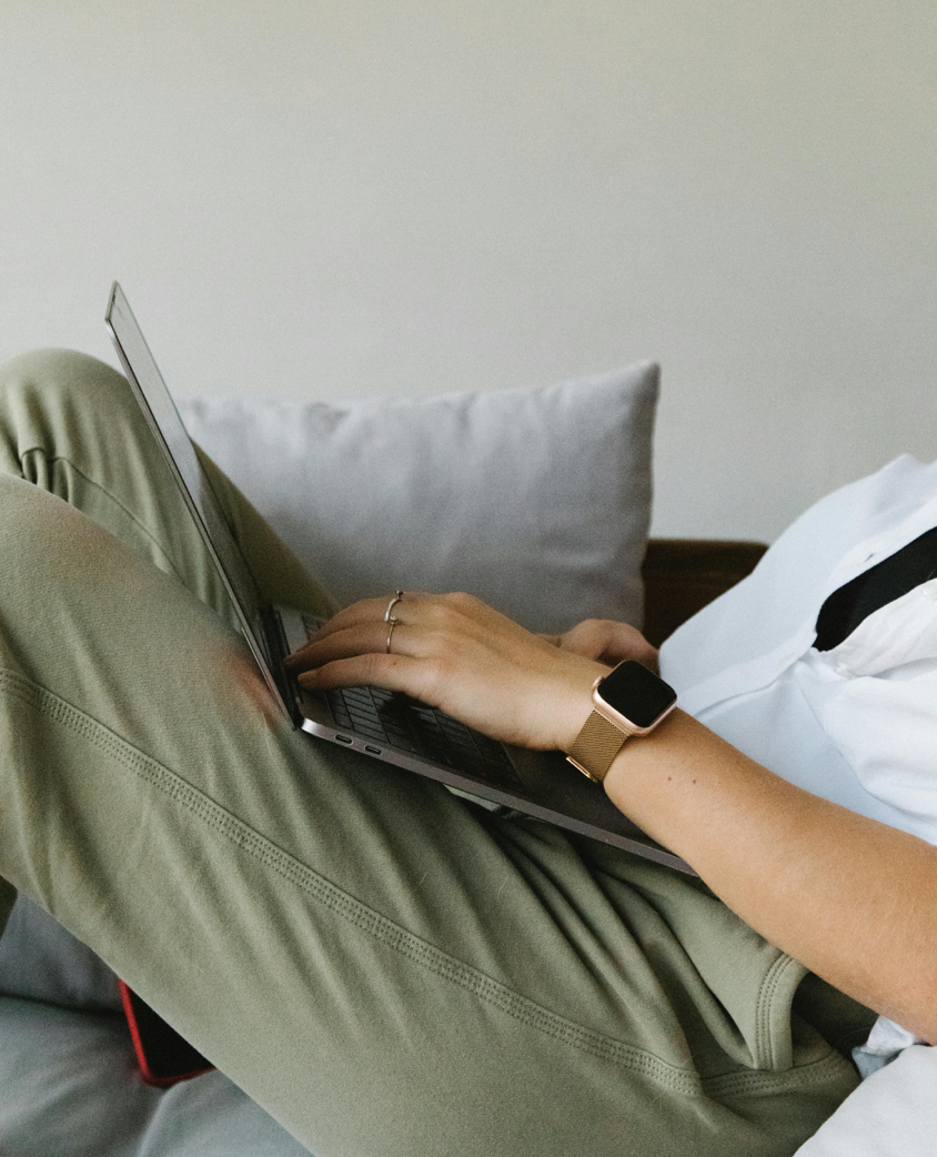 Side view of crop unrecognizable female freelancer in casual clothes and smartwatch lying on comfortable sofa and working remotely on laptop